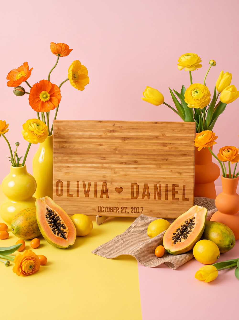 Bamboo cutting board engraved with two bold names, a heart and date, styled with yellow flowers on pink.