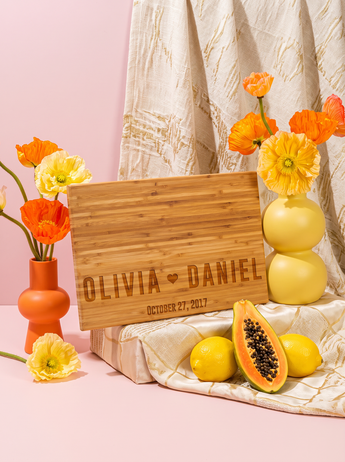 Angled view of a bamboo cutting board engraved with two bold names, a heart and date, beside yellow and orange flowers.