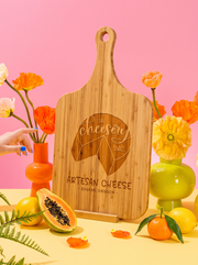 Custom logo engraved on a bamboo handle cutting board, styled with colorful flowers against a pink background.