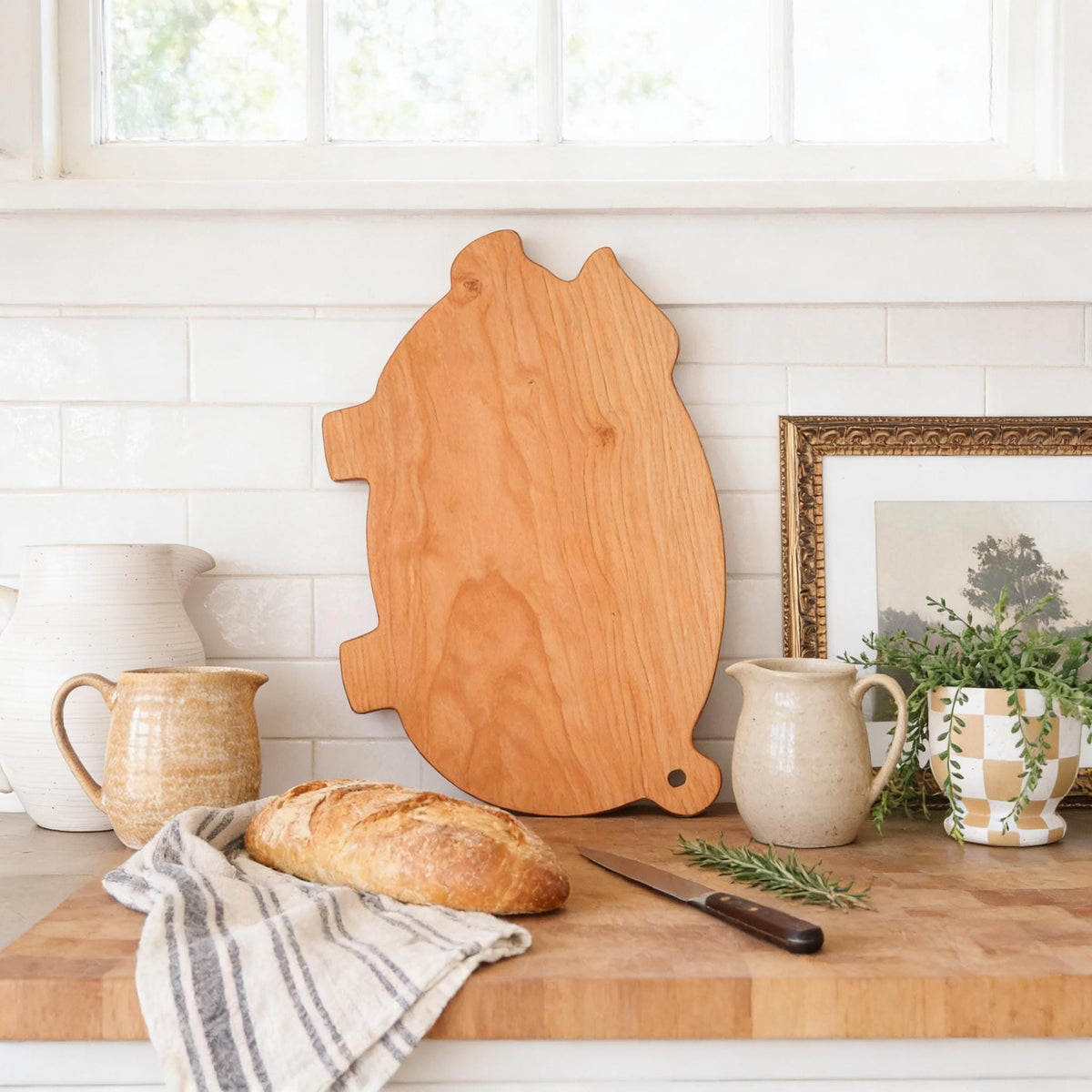 a wooden cutting board, a loaf of bread, a pitcher, a vase, and a potted plant arranged on a wooden countertop.