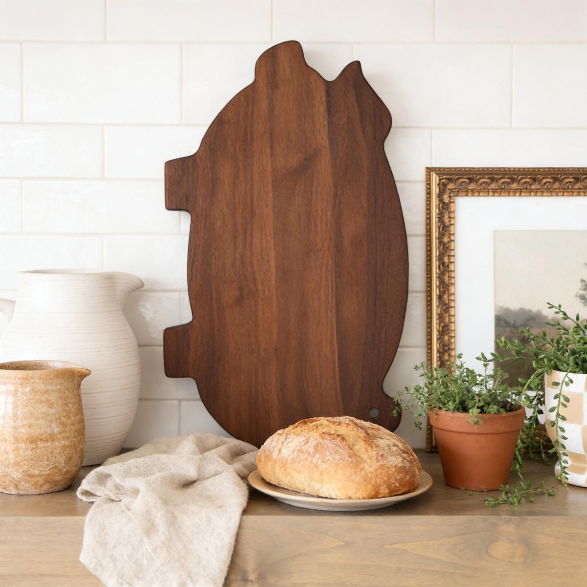 a wooden cutting board, a plate of bread, a white vase, a potted plant, and a framed picture on a wooden shelf.