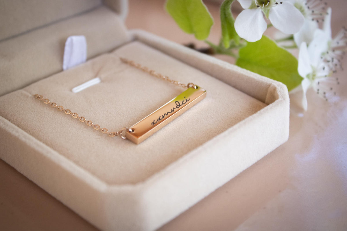 A gold-colored necklace with a rectangular pendant is displayed in a beige jewelry box, accompanied by a bouquet of white flowers.