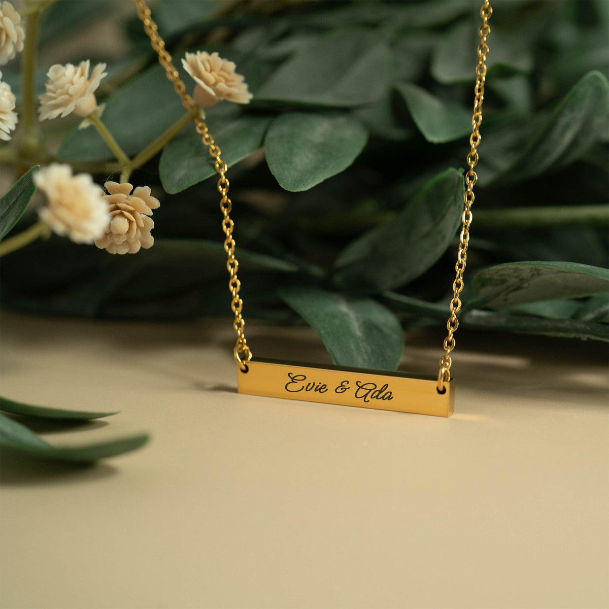 A gold-colored necklace with a rectangular pendant engraved with the names "Eve & Ada". The necklace is displayed against a backdrop of green leaves and white flowers.