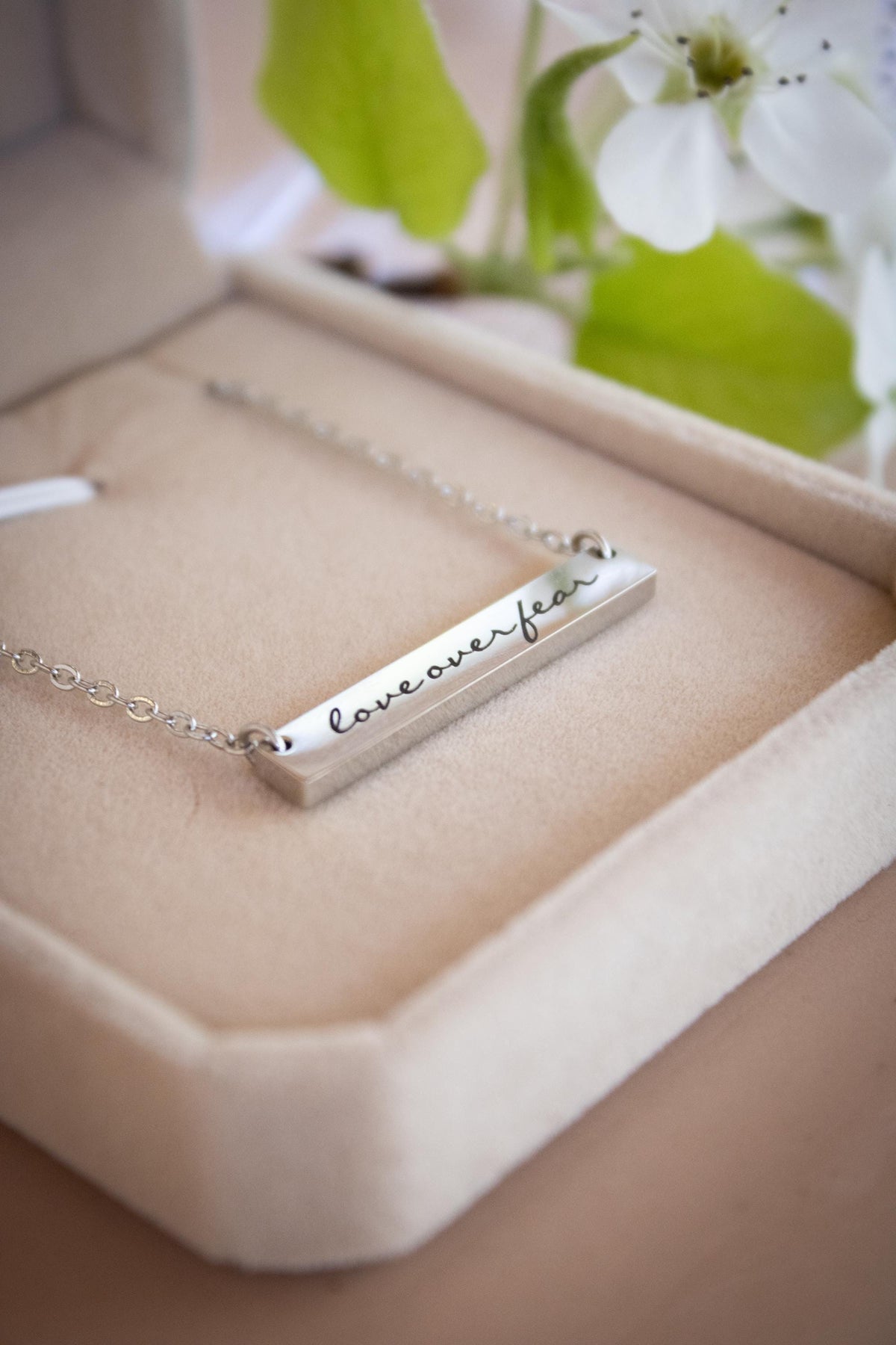 A necklace with a rectangular pendant that says "loveover fear" engraved on it, placed in a white jewelry box.