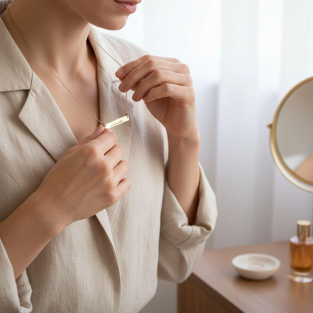 A woman in a beige blazer is adjusting a necklace with a pendant in front of a mirror.