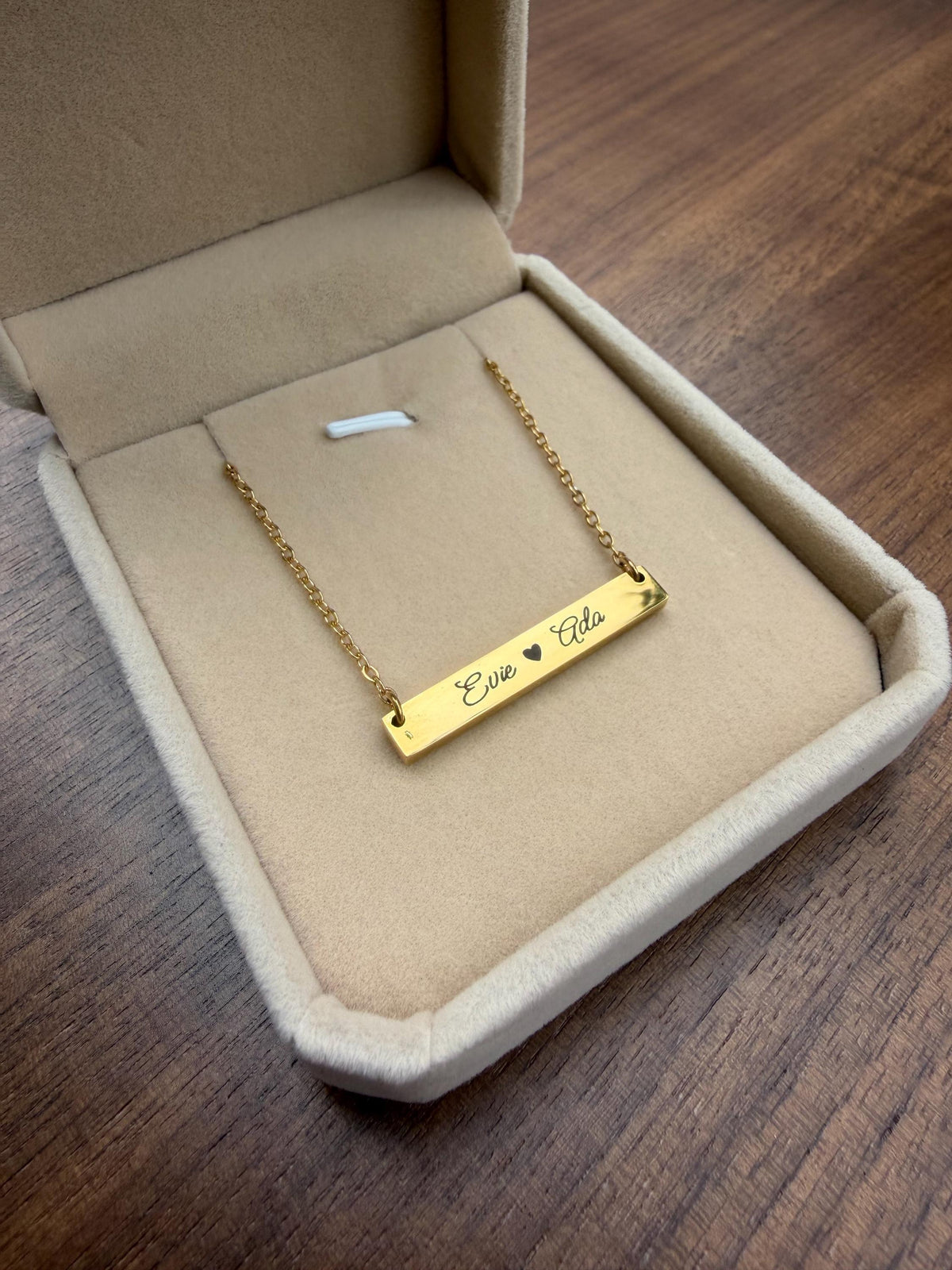 May include: A gold-colored bar necklace with the names "Evie" and "Ada" engraved on it, separated by a heart symbol. The necklace is displayed inside a beige jewelry box with a velvet lining. The box is open, revealing the necklace.