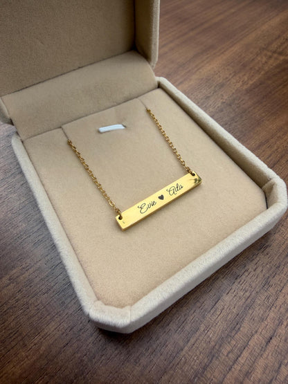 May include: A gold-colored bar necklace with the names "Evie" and "Ada" engraved on it, separated by a heart symbol. The necklace is displayed inside a beige jewelry box with a velvet lining. The box is open, revealing the necklace.