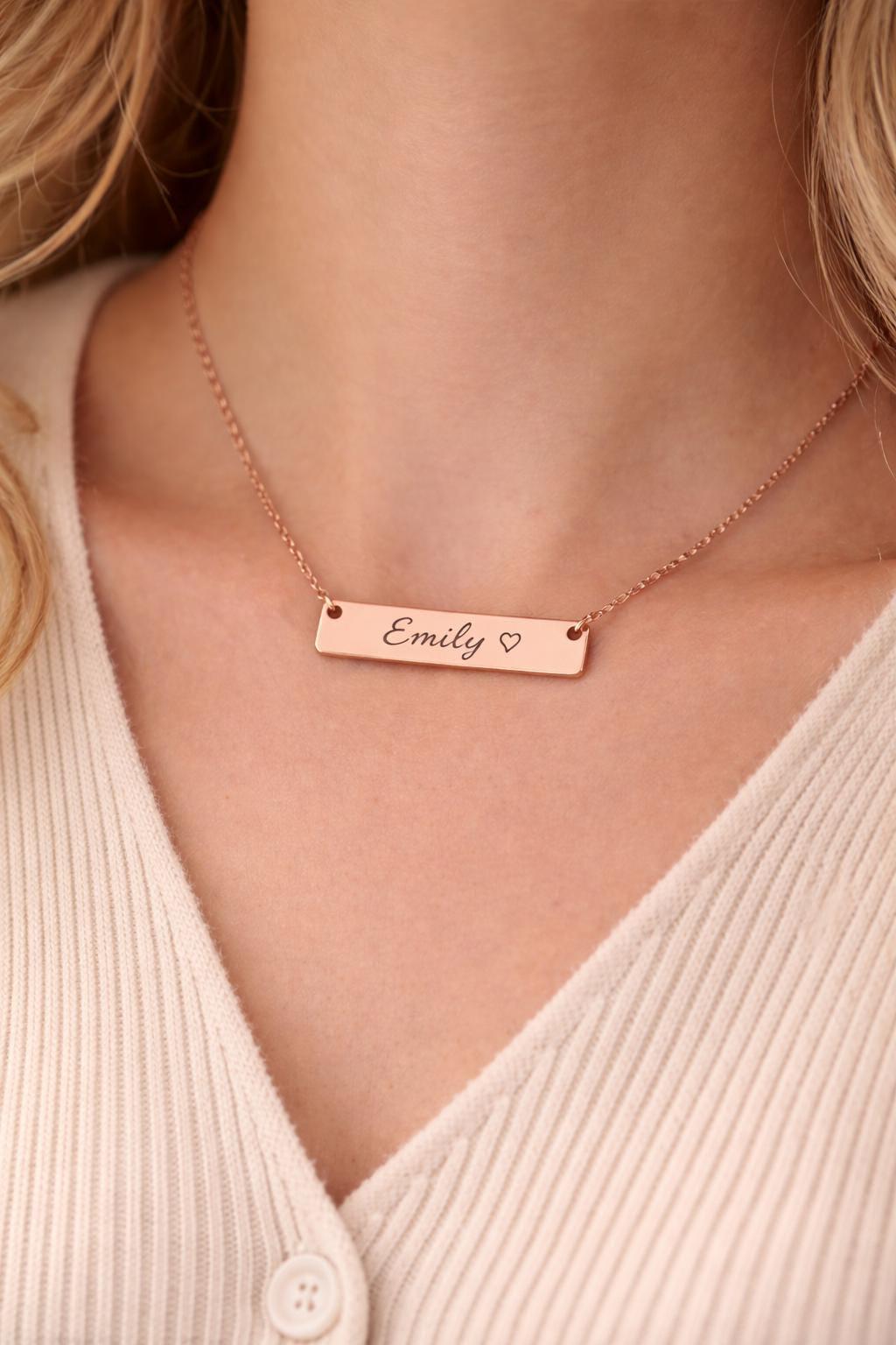 A close-up of a woman wearing a rose gold bar necklace with the name 'Emily' and a small heart engraved on it, paired with a cream ribbed cardigan.