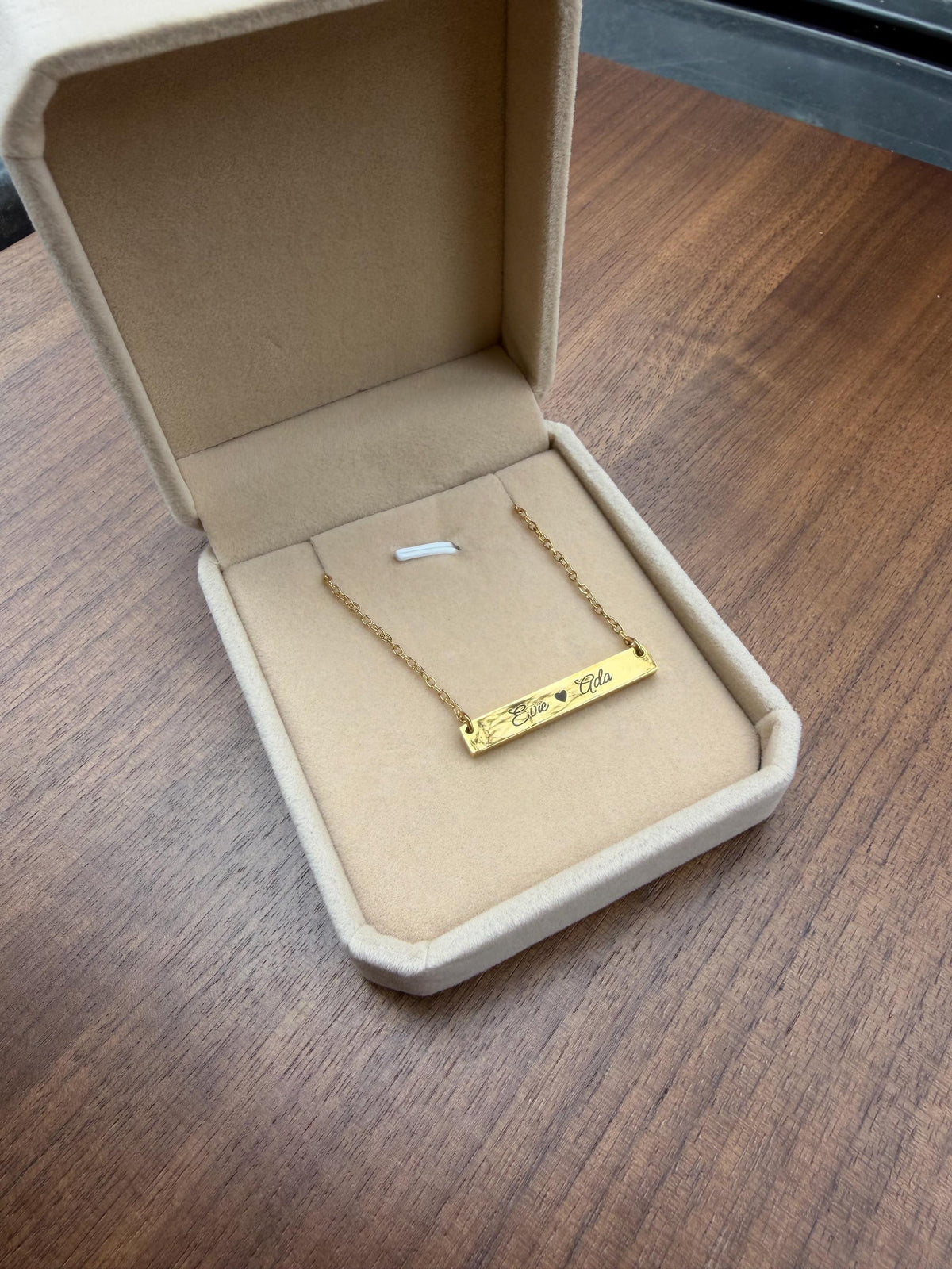A gold bar necklace with engraved text resting inside an open beige jewelry box on a wooden table.