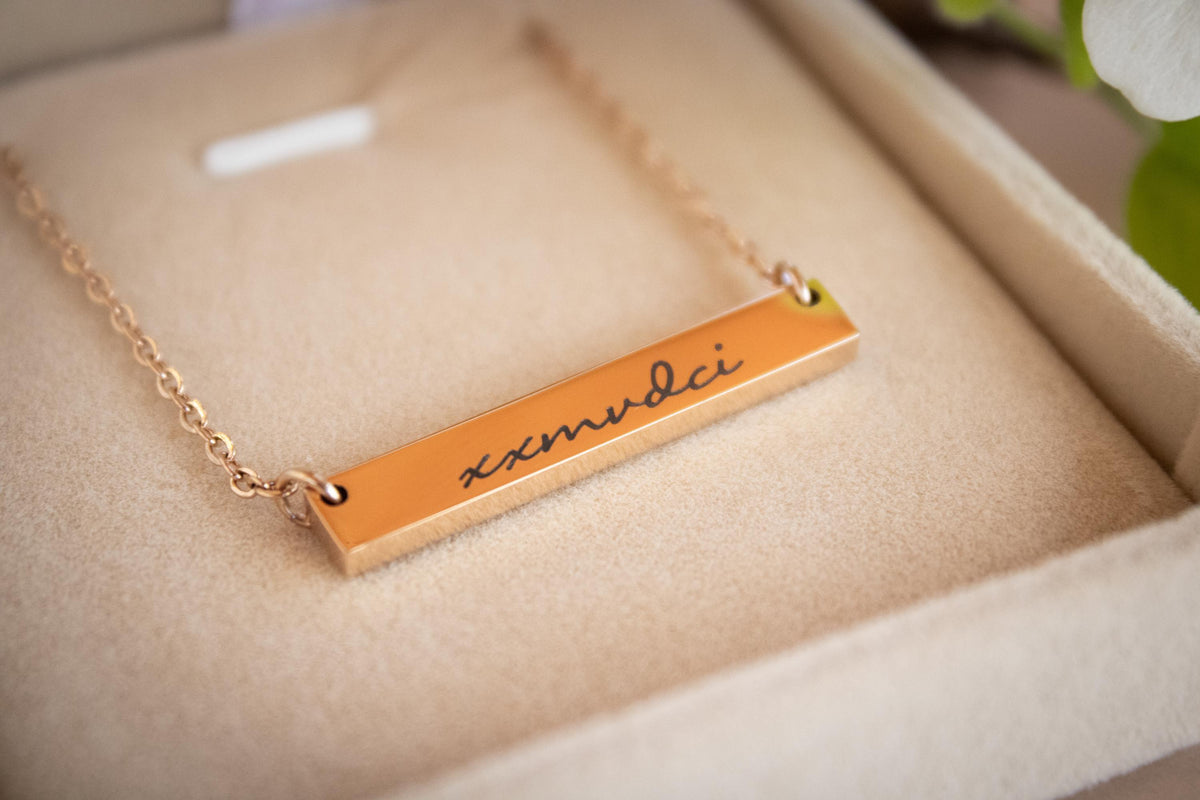 A rose gold bar necklace engraved with the name 'emudci' in black script, resting inside a beige jewelry box.