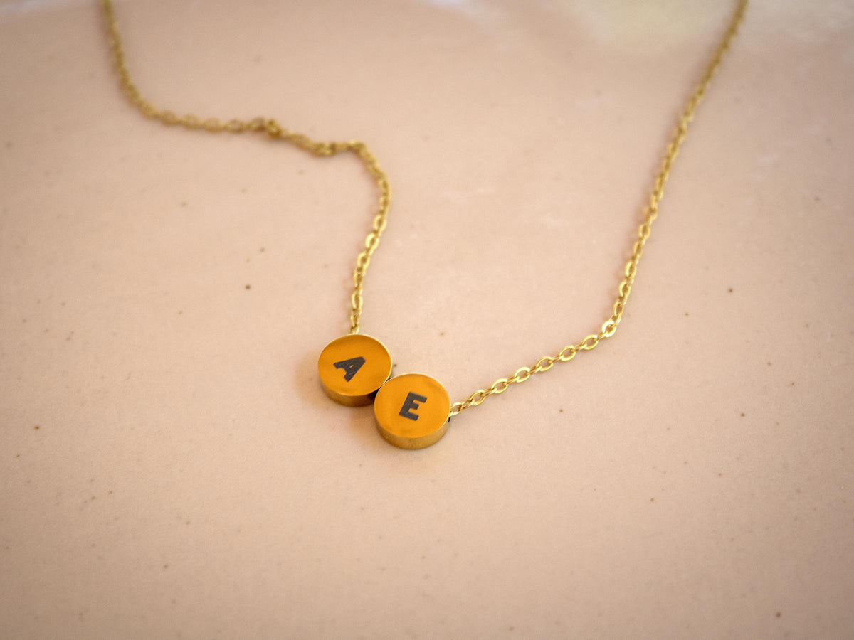 A gold chain necklace with two round yellow-orange initial pendants spelling 'AE', resting on a light beige textured surface.