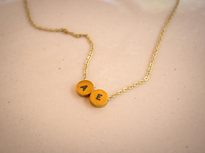 A gold chain necklace with two round yellow-orange initial pendants spelling 'AE', resting on a light beige textured surface.