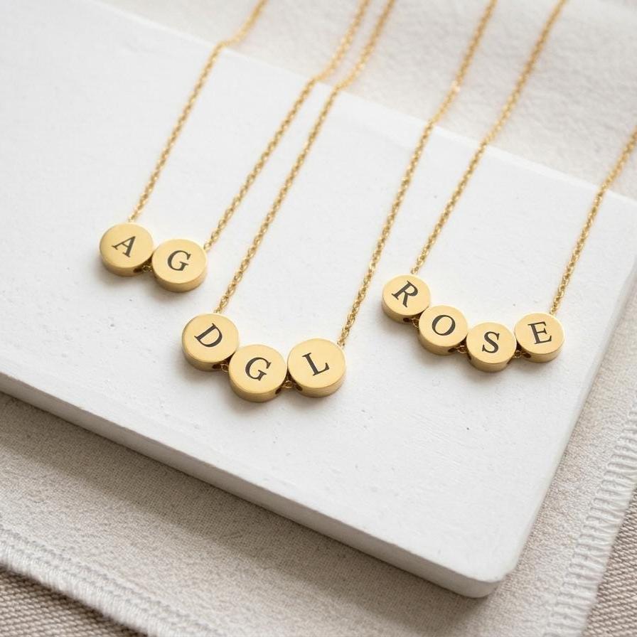 Three gold initial necklaces with round letter beads spelling AG, DGL, and ROSE, arranged on a white rectangular tray over a beige fabric background.