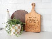 Holly Mixing Bowl edited mockup on a warm-toned wooden handled board with Nana’s Christmas Bakery artwork, beside a plant against white tile.