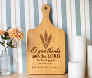 Generated mockup of a bamboo paddle board with wheat, O give thanks scripture, Psalm 107:1 and The Bennett Family, beside flowers and candles.