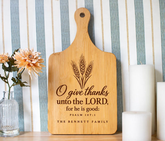Generated mockup of a bamboo paddle board with wheat, O give thanks scripture, Psalm 107:1 and The Bennett Family, beside flowers and candles.