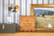 Edited PSD composite mockup of a scalloped square board with O give thanks, Psalm 107:1 and The Bennett Family, beside flowers and framed art.