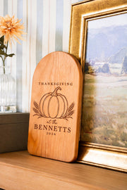 Pumpkin wheat Thanksgiving engraving mockup on an arched board, personalized BENNETTS 2026, beside a framed landscape.