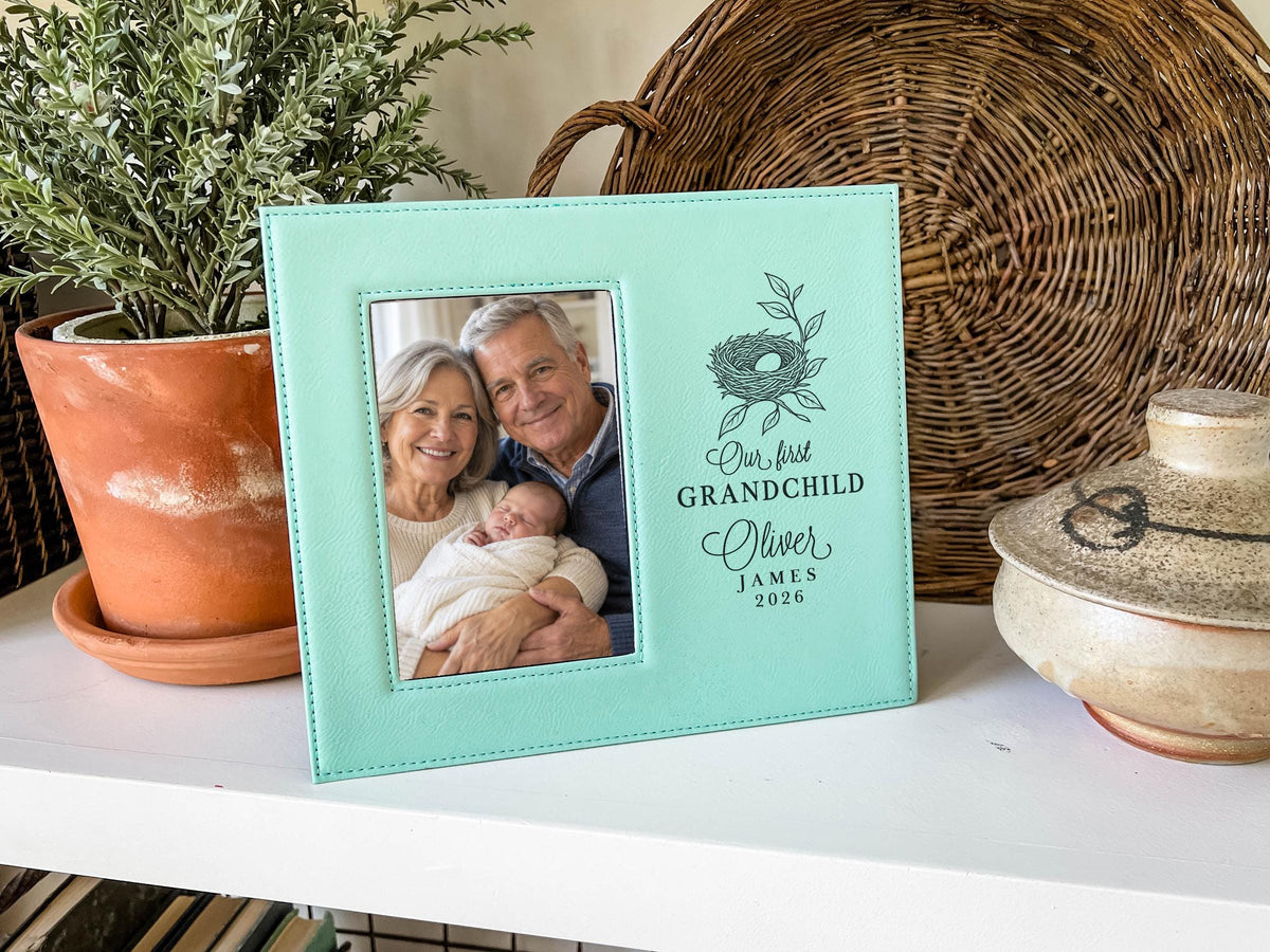 Teal-colored frame beside a potted plant and woven tray, showing a nest design, “Our first GRANDCHILD,” sample Oliver / JAMES / 2026, and an illustrative portrait of grandparents holding a newborn.