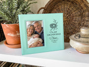Teal-colored frame beside a potted plant and woven tray, showing a nest design, “Our first GRANDCHILD,” sample Oliver / JAMES / 2026, and an illustrative portrait of grandparents holding a newborn.