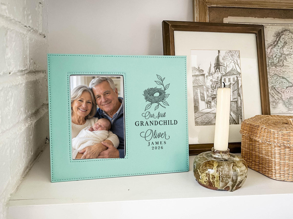 Teal-colored frame on a shelf beside a candle, showing a nest design, “Our first GRANDCHILD,” sample Oliver / JAMES / 2026, and an illustrative portrait of grandparents holding a newborn.