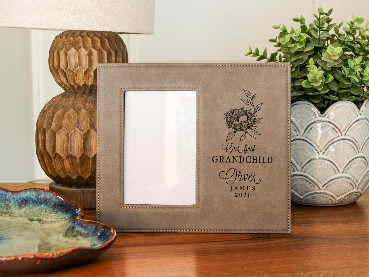 Taupe-colored frame beside a lamp and plant, with an empty portrait opening and nest engraving reading “Our first GRANDCHILD,” with sample Oliver / JAMES / 2026. No photograph is inserted.