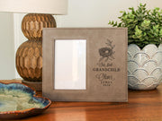 Taupe-colored frame beside a lamp and plant, with an empty portrait opening and nest engraving reading “Our first GRANDCHILD,” with sample Oliver / JAMES / 2026. No photograph is inserted.
