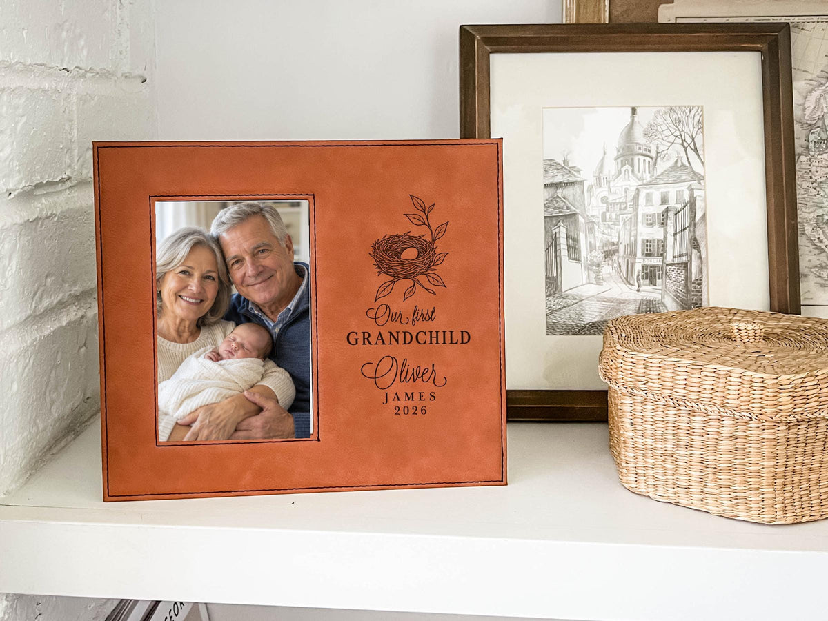 Brown-colored frame on a shelf beside a woven basket, showing a nest design, “Our first GRANDCHILD,” sample Oliver / JAMES / 2026, and an illustrative portrait of grandparents holding a newborn.