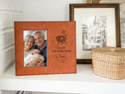 Brown-colored frame on a shelf beside a woven basket, showing a nest design, “Our first GRANDCHILD,” sample Oliver / JAMES / 2026, and an illustrative portrait of grandparents holding a newborn.