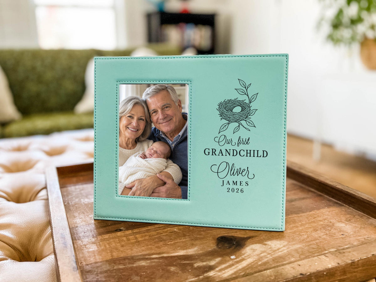 Teal-colored frame on a tray in a living room, showing a nest design, “Our first GRANDCHILD,” sample Oliver / JAMES / 2026, and an illustrative portrait of grandparents holding a newborn.