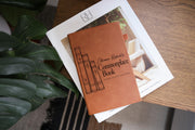 Angled edited mockup of a brown Commonplace Book journal resting on a white book beside green leaves; three illustrated book spines, sample name Eleanor Robert’s and Notes & Reflections.