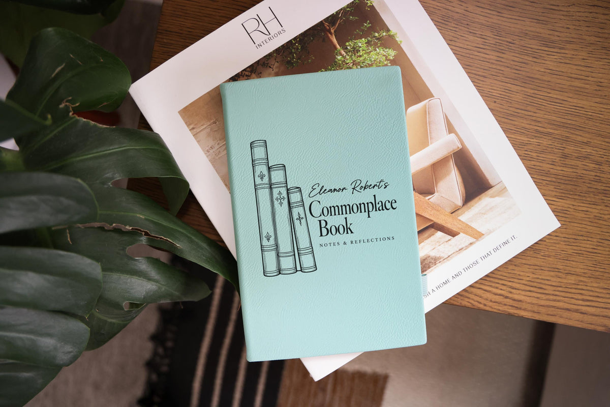 Edited mockup of a teal journal resting on an interiors book beside green leaves, showing the sample name Eleanor Robert’s, Commonplace Book and Notes & Reflections.