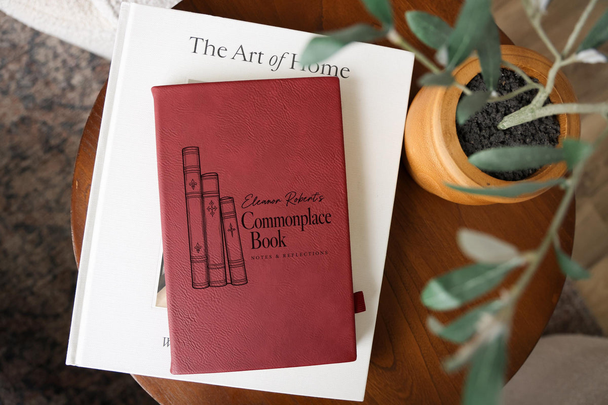 Edited mockup of a rose-red Commonplace Book journal on The Art of Home beside a potted plant, with three illustrated book spines and sample name Eleanor Robert’s.