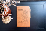 Chestnut Garden Hydrangea journal mockup on a dark table with a matching pen and dried flowers. Cover shows a hydrangea, Garden Observations, Julia Bennett, and Field Notes.