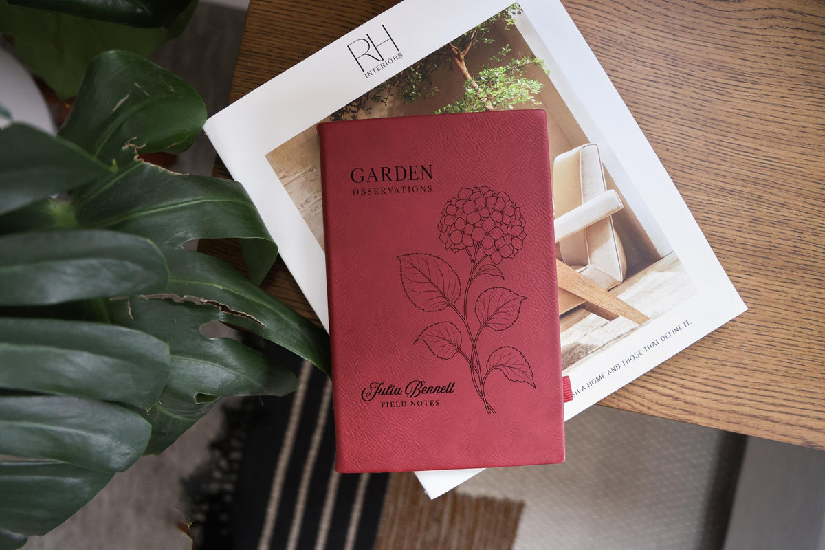Rose Garden Hydrangea journal mockup on a book beside green leaves and a wood table. Cover shows a hydrangea, Garden Observations, Julia Bennett, and Field Notes.