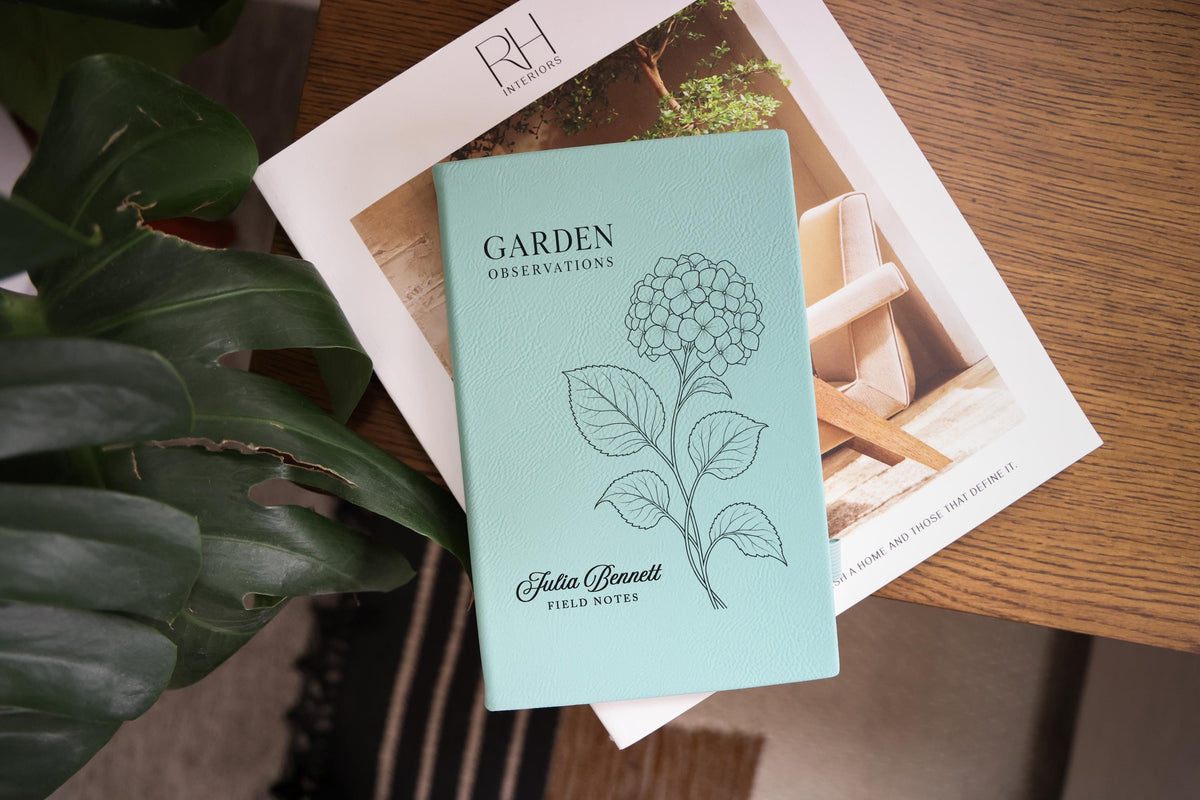 Teal Garden Hydrangea journal mockup on a book beside green leaves and a wood table. Cover shows a hydrangea, Garden Observations, Julia Bennett, and Field Notes.
