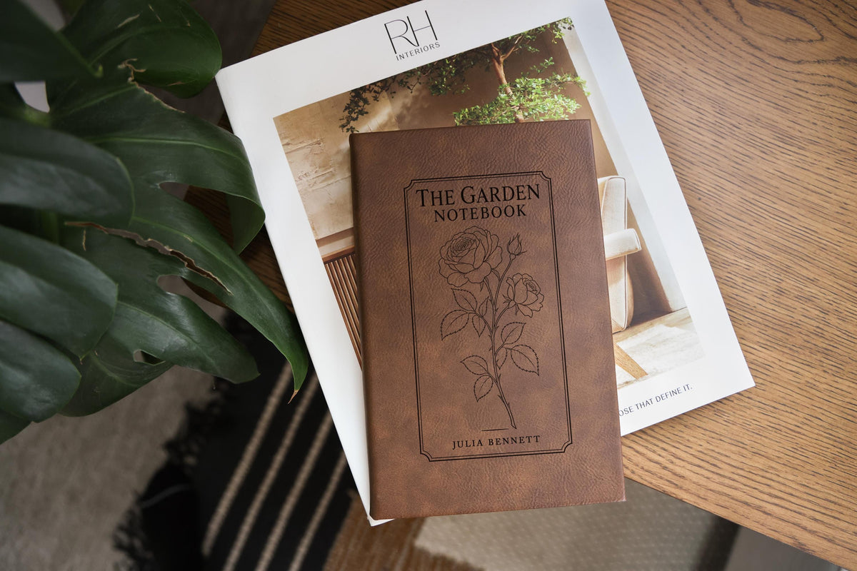 Brown The Garden journal mockup on a book beside broad green leaves and a wood table. Cover shows a rose stem, a fine border, The Garden Notebook, and the sample name Julia Bennett.