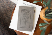 Gray The Garden journal mockup on a book beside a potted plant. Cover shows a rose stem, a fine border, The Garden Notebook, and the sample name Julia Bennett.