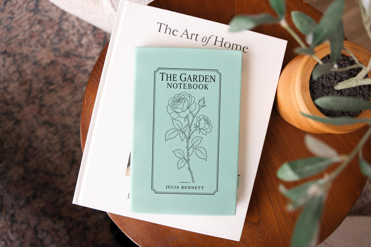Teal The Garden journal mockup on a book beside a potted plant. Cover shows a rose stem, a fine border, The Garden Notebook, and the sample name Julia Bennett.