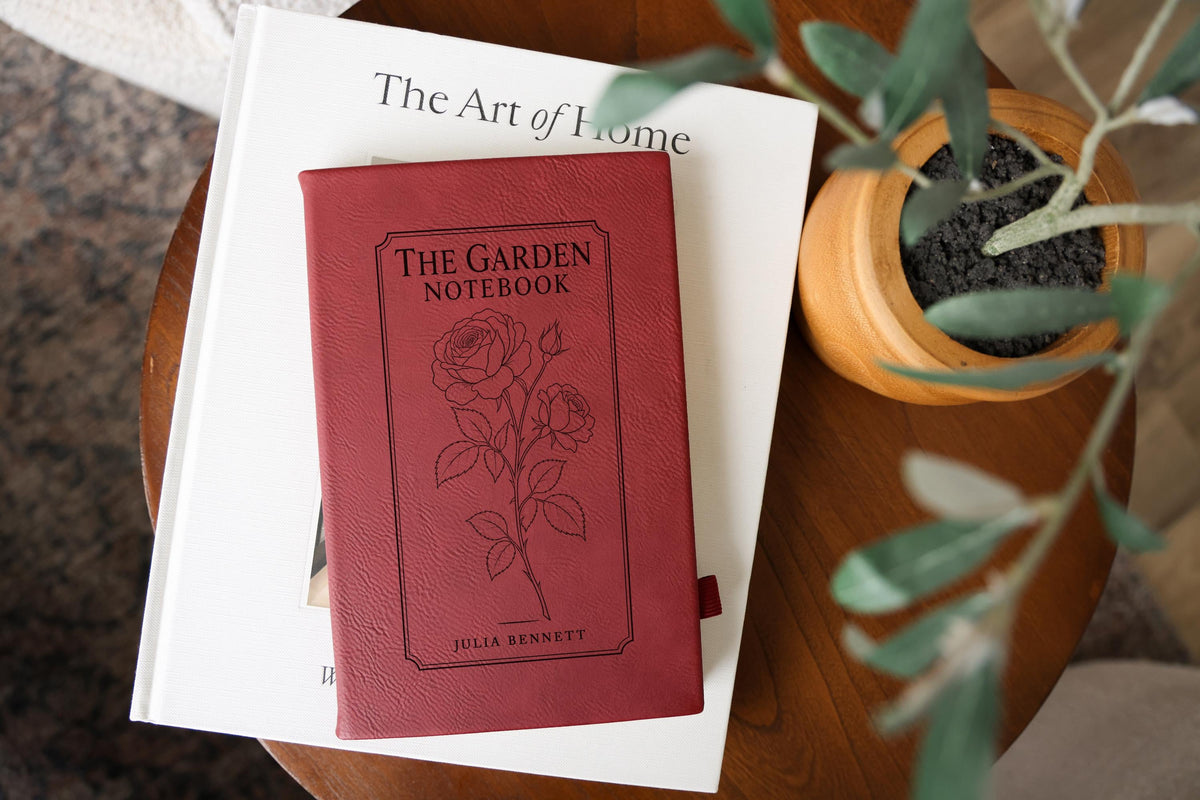 Rose The Garden journal mockup on a book beside a potted plant. Cover shows a rose stem, a fine border, The Garden Notebook, and the sample name Julia Bennett.