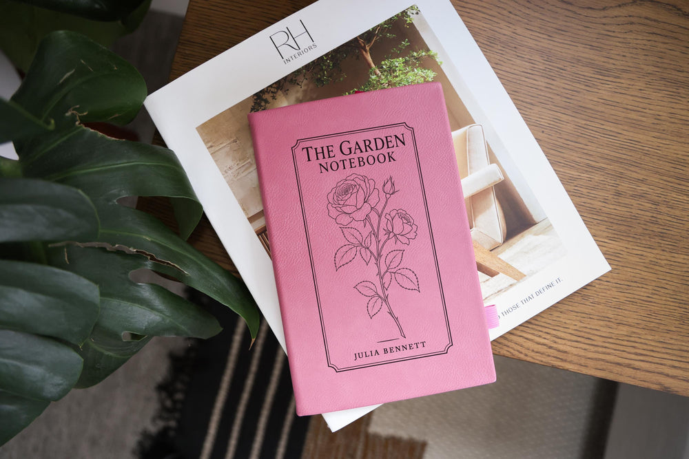Pink The Garden journal mockup on a book beside broad green leaves and a wood table. Cover shows a rose stem, a fine border, The Garden Notebook, and the sample name Julia Bennett.