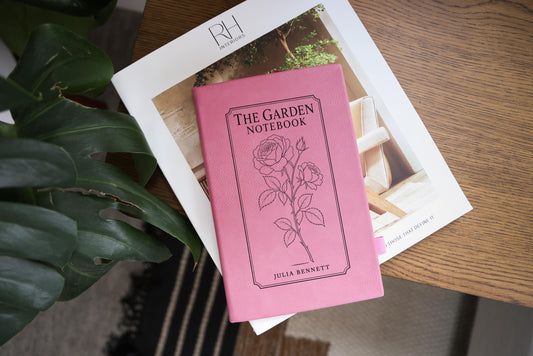 Pink The Garden journal mockup on a book beside broad green leaves and a wood table. Cover shows a rose stem, a fine border, The Garden Notebook, and the sample name Julia Bennett.