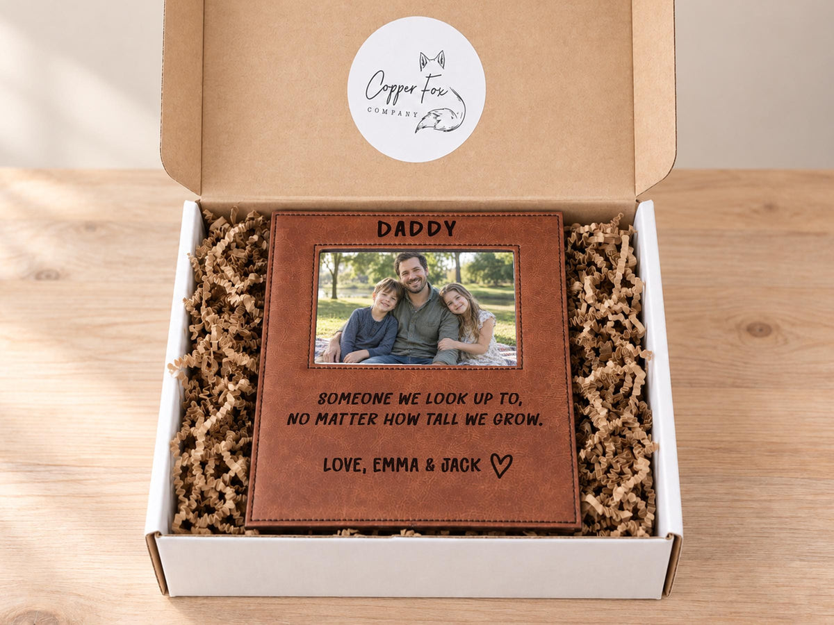 Edited mockup of the tall we grow Daddy frame inside an open gift box with crinkle paper, with a sample dad-and-children photo, engraved saying, Love, Emma & Jack signature and outline heart.