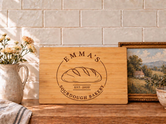 Edited mockup of a bamboo rectangle cutting board with Emma’s Sourdough Bakery loaf emblem and EST. 2020, styled on a kitchen counter.