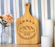 Edited mockup of a bamboo board with a handle, showing Emma’s Sourdough Bakery engraving beside flowers and candles.
