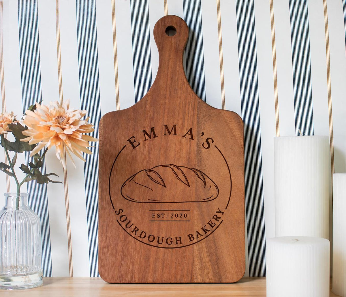 Edited mockup of a walnut board with a handle and Emma’s Sourdough Bakery loaf emblem, beside flowers and candles.