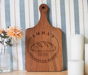 Edited mockup of a walnut board with a handle and Emma’s Sourdough Bakery loaf emblem, beside flowers and candles.