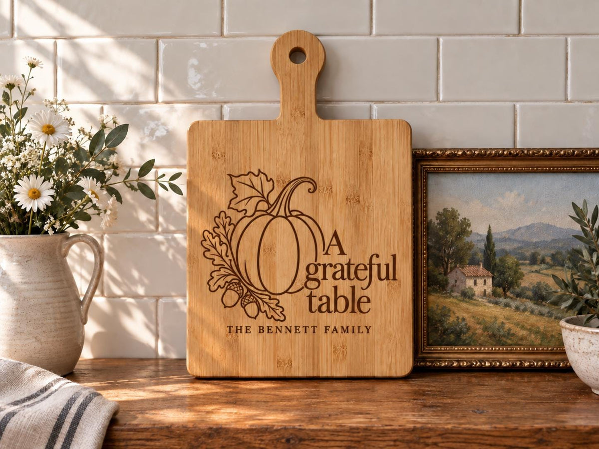 Grateful Table pumpkin and Bennett family engraving mockup on a bamboo paddle board beside flowers and a landscape painting.