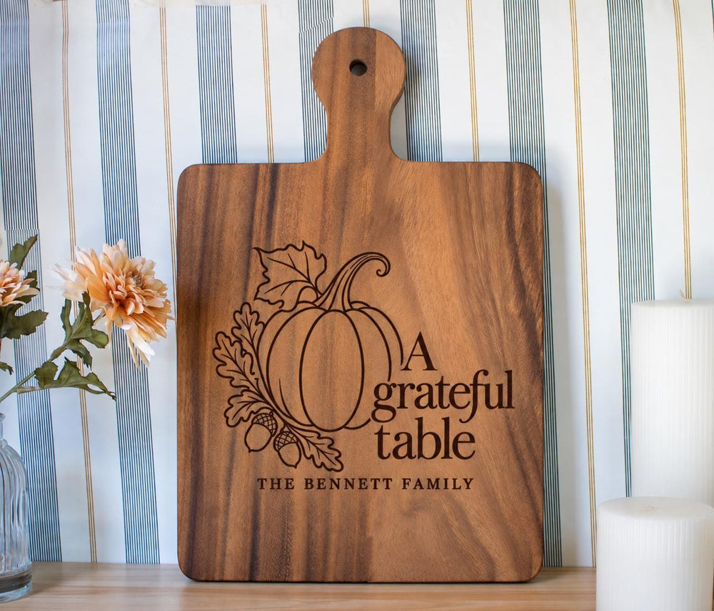 Grateful Table pumpkin, oak leaves and Bennett family engraving mockup on a walnut paddle board beside flowers and candles.