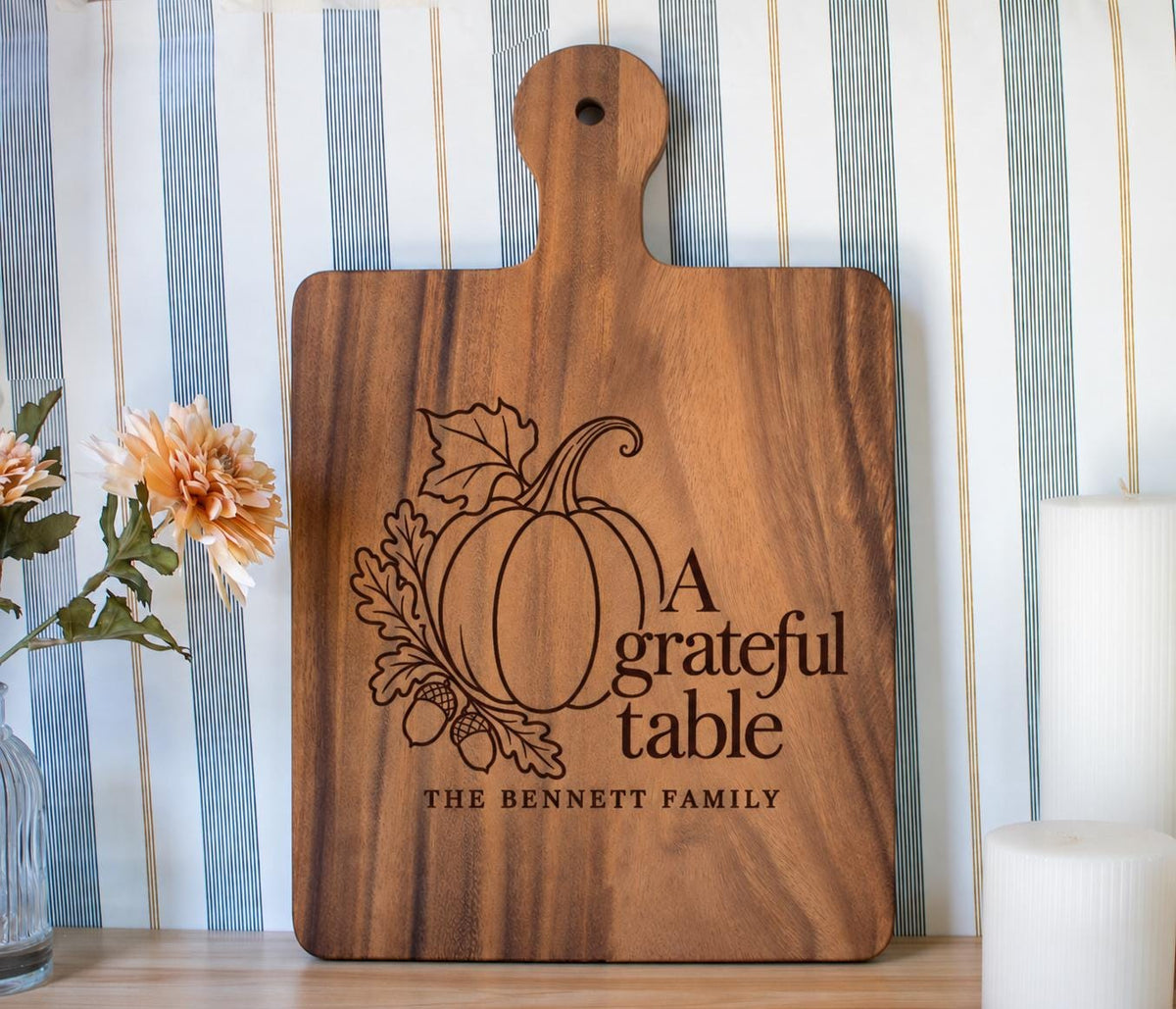 Grateful Table pumpkin, oak leaves and Bennett family engraving mockup on a walnut paddle board beside flowers and candles.